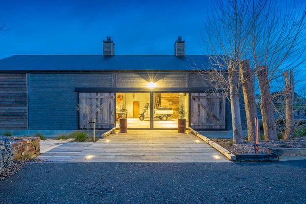 Twilight exterior view of a single-storey barn-like building with dark horizontal timber cladding, a low pitched roof, and a warmly illuminated central entrance. Large weathered timber barn doors are opened on either side of the glazed entry, with a timber boardwalk leading across a gravel forecourt. Bare trees and low stone landscaping frame the approach.