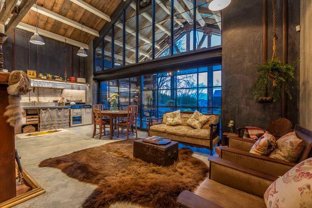 A rustic industrial open-plan living area with a compact kitchen and dining zone. The space features exposed timber roof framing, dark metal-framed floor-to-ceiling glazing, textured dark walls, timber furniture, a vintage-style sofa and armchairs, pendant lighting, and layered natural-fibre rugs. A large glazed upper void creates an airy, loft-like character.