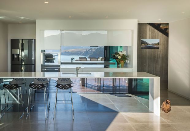 Bright contemporary open-plan kitchen with a large white island and integrated seating, dark appliances, reflective cabinetry, and expansive glazing overlooking a mountainous landscape. A dark timber feature wall with framed views adds contrast, while polished concrete or large-format grey flooring and recessed ceiling lights create a clean, modern feel. A cat is visible at the edge of the room.