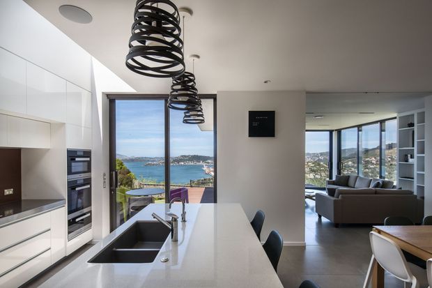 Contemporary open-plan kitchen and living area with a large white island, integrated appliances, dark sink and tapware, and sculptural black spiral pendant lights. Floor-to-ceiling glazing provides expansive waterfront views, while the adjacent living and dining zones are visible beyond.