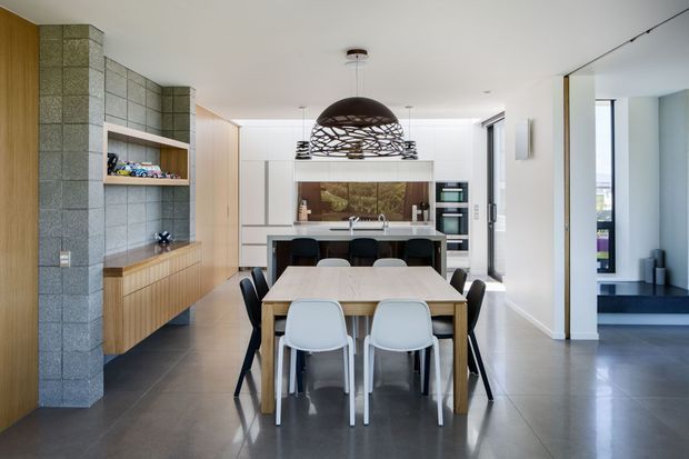 Open-plan contemporary dining and kitchen area with a large timber dining table and mixed seating in the foreground. A sculptural woven pendant light hangs above the table, while the rear kitchen features pale built-in cabinetry, a dark island, integrated appliances and a timber-framed splashback. The space has polished dark flooring, a grey masonry feature wall with timber built-in shelving, and an opening to a lower-level or adjacent living area on the right.