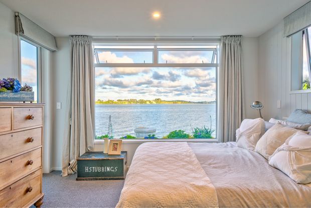 Bright, calm bedroom overlooking a broad body of water through a large picture window. The room features a neatly made bed with pale quilted bedding, light curtains, white painted walls with vertical panel detailing, a timber chest of drawers, bedside lamp, and soft grey carpet. The waterfront view and abundant natural light create an airy coastal atmosphere.