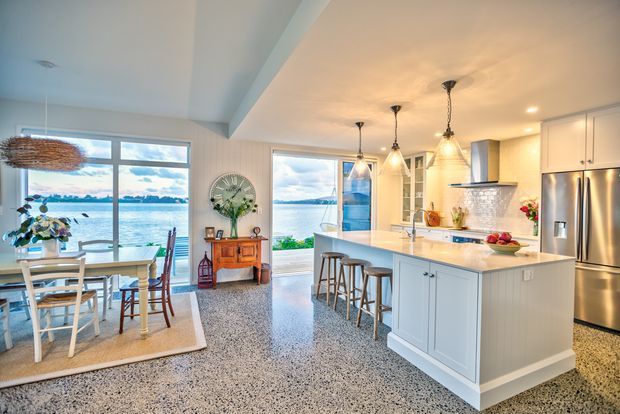 Bright open-plan kitchen and dining area with a large white island, pendant lights, stainless-steel appliances, shaker-style cabinetry, and a terrazzo-like speckled floor. The adjacent dining space has large windows overlooking a coastal waterfront, with a small vintage console and natural woven lighting adding character.