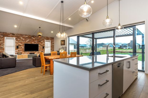Open-plan contemporary kitchen and living area with a large white island, dark stone benchtop, integrated dishwasher, pendant lighting, timber flooring, exposed brick feature wall, dining table, and lounge seating. Full-height glazed doors open to an outdoor area and distant residential landscape.