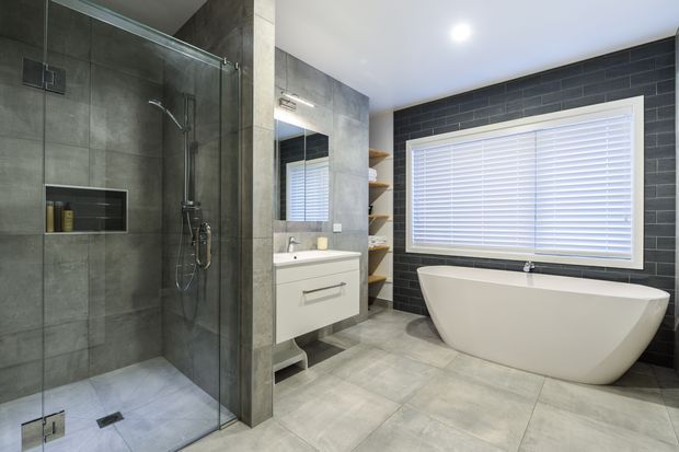 A contemporary bathroom with a large frameless glass walk-in shower, wall-mounted rain shower and handheld fixture, floating white vanity, freestanding white bathtub, and open timber shelving. Large-format grey floor and wall tiles contrast with a dark charcoal brick-pattern feature wall around the window. The room is bright, clean, and refined, with white plantation shutters.