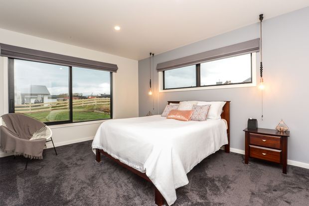 A bright, contemporary bedroom with a large bed dressed in white linen, a dark timber headboard and matching bedside table, grey carpet, pale grey walls, and expansive black-framed windows overlooking a rural landscape. Hanging pendant lights and a soft upholstered chair complete the room.