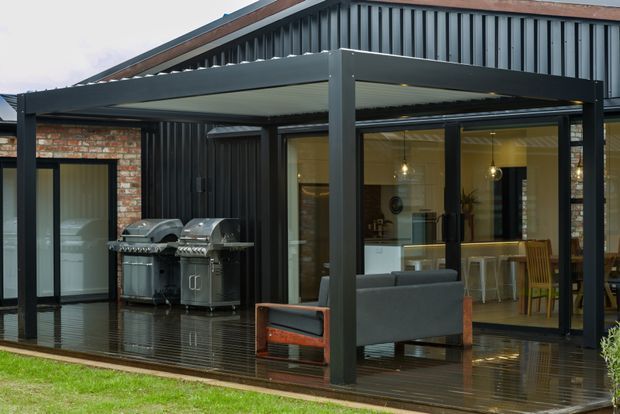 Exterior view of a contemporary single-storey home extension or covered outdoor entertaining area. The structure combines dark corrugated metal cladding, exposed brick walls, extensive glazing, and a flat-roofed steel-framed canopy over a timber deck. Outdoor grills, seating, and an interior kitchen/living area are visible through the glazing.