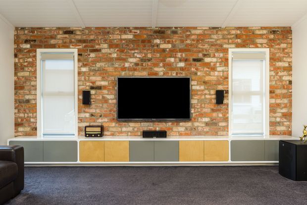 A contemporary media wall with a wall-mounted television, speakers, and low built-in cabinetry set against an exposed red brick feature wall. Two tall windows flank the television, while a white panelled ceiling, grey carpet, and dark seating complete the room.