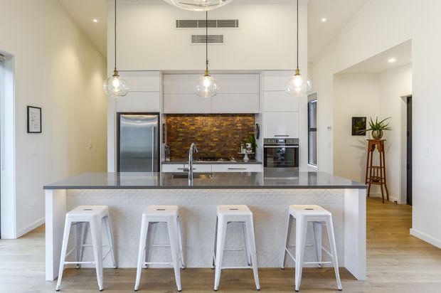 Bright contemporary open-plan kitchen featuring a large central island with four white stools, white cabinetry, stainless steel appliances, a reflective brown mosaic tile splashback, pendant lights, and pale timber flooring. A hallway and small styled nook are visible to the right.