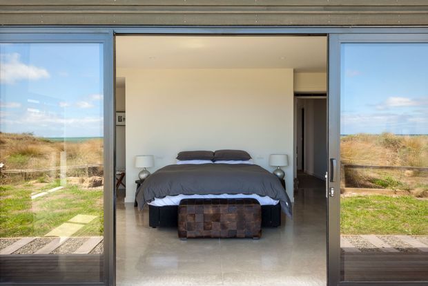 A contemporary bedroom viewed through wide sliding glass doors, with a neatly made bed, bedside lamps, and a low upholstered bench. The room opens directly onto a landscaped outdoor area, with expansive glazing and a simple neutral palette.