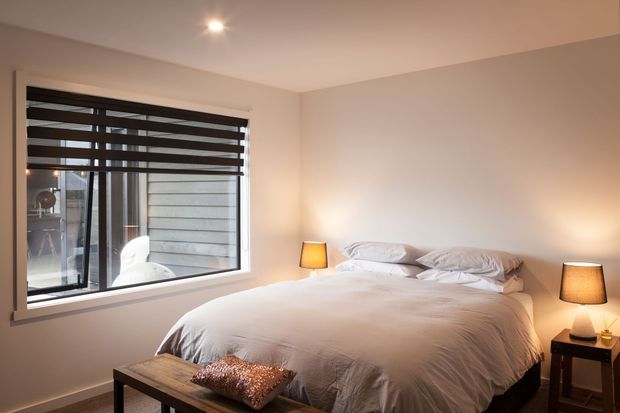 A warm, simply furnished bedroom with a large window, layered dark roller blinds, a low bed dressed in white linen, matching bedside lamps, and a wooden bench at the foot of the bed. The neutral palette and soft lighting create a calm, inviting atmosphere.