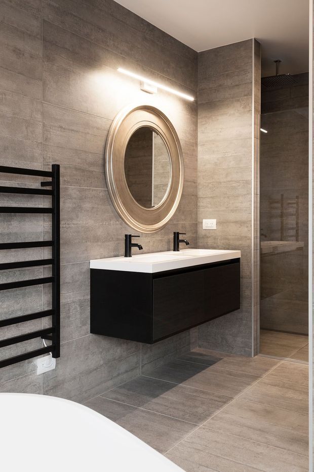 Modern bathroom interior featuring large-format grey concrete-look wall and floor tiles, a floating dark vanity with white double basins and black tapware, a round metallic-framed mirror, a black heated towel rail, and a frameless glass shower enclosure. The palette is contemporary, neutral, and refined.