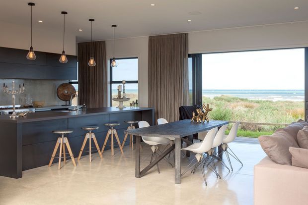 Open-plan coastal kitchen, dining and living area with dark streamlined cabinetry, a large island with timber stools, a dark dining table with white sculptural chairs, pendant lighting, polished light flooring and expansive glazing overlooking dunes and the ocean.