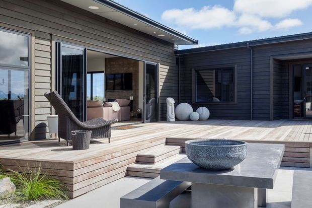 Contemporary residential courtyard and deck with dark horizontal timber cladding, broad timber decking, large glazed openings, and stepped platform seating. A sculptural outdoor table with a decorative stone bowl occupies the foreground, while an open living area is visible through the doors. The composition is bright, clean, and minimalist with strong indoor-outdoor connection.