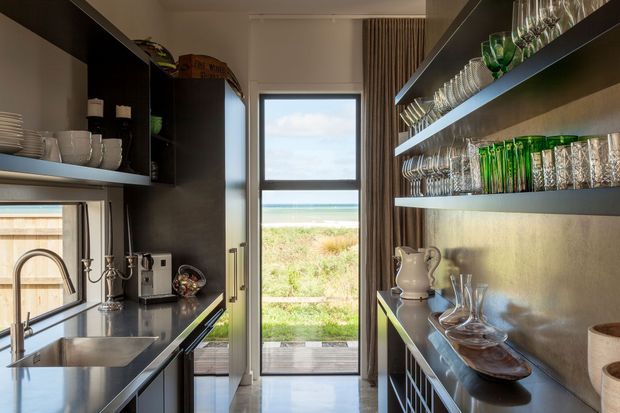 A narrow, sophisticated galley kitchen and drinks service area with dark cabinetry, stainless-steel benchtops, open shelving filled with glassware, a sink and tap on the left, and a glazed door at the rear opening toward a coastal landscape. The space has warm concealed lighting, full-height curtains, and a polished contemporary fit-out.