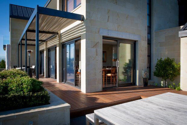 Contemporary courtyard-side view of a light stone-clad residence with extensive dark-framed glazing and sliding doors opening onto a raised timber deck. A slatted pergola provides shade over the outdoor dining area, with landscaped planters and a large outdoor table in the foreground. Interior dining furnishings are visible through the open glazing.
