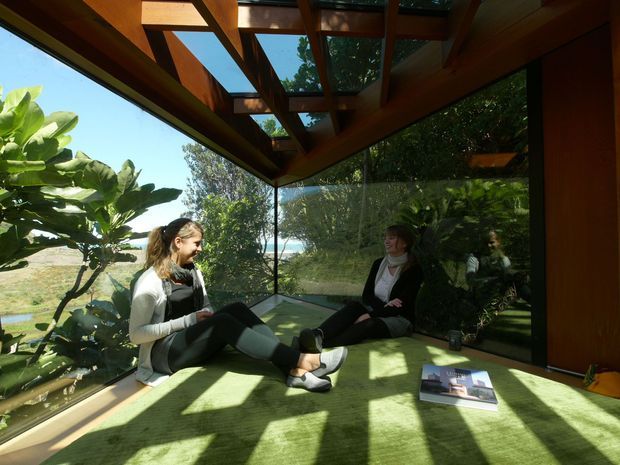 A compact, glass-walled sunroom or glazed retreat with a dramatic exposed timber roof structure and extensive floor-to-ceiling glazing. Two people sit on a green carpeted floor, with lush planting and a distant water or coastal landscape visible outside. Warm timber framing, strong sunlight, and reflected greenery create an intimate indoor-outdoor atmosphere.