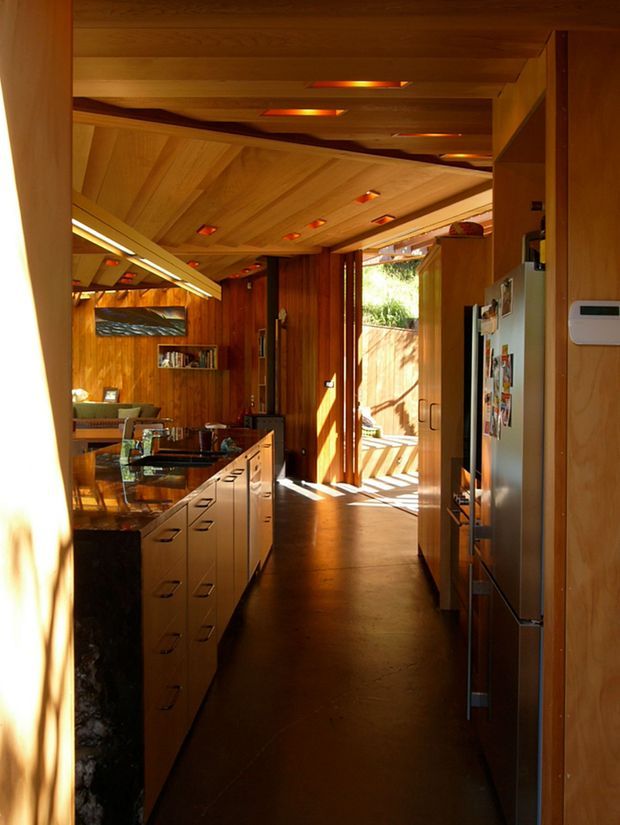 Warm, narrow open-plan residential interior viewed from a kitchen passage. The space features extensive honey-toned timber wall and ceiling panelling, exposed timber ceiling beams, integrated recessed lighting, a long kitchen island with pale built-in drawers and a dark stone benchtop, and a stainless-steel refrigerator. A dining or living area is visible beyond, with strong sunlight entering through an open glazed doorway to an outdoor area.