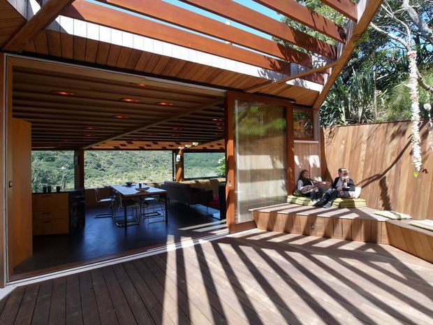 A warm timber-clad indoor-outdoor living and dining area opens onto a spacious timber deck. A pergola with broad timber beams casts strong linear shadows across the deck, while built-in bench seating runs along a timber privacy wall. Through the wide glazed opening, a dining table, kitchen joinery and lounge seating are visible, with leafy hills beyond.