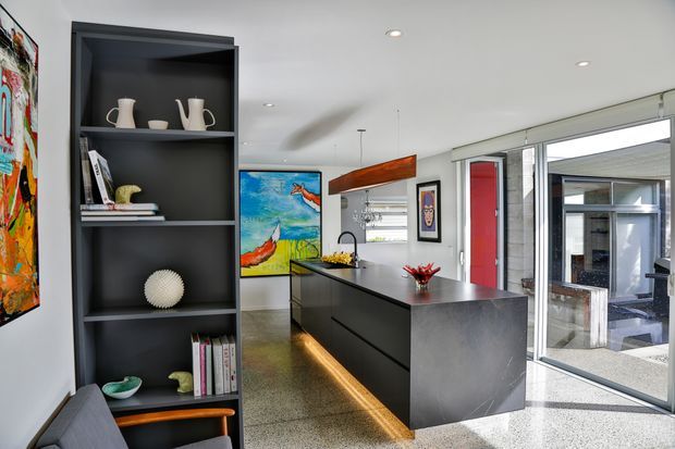 Bright contemporary kitchen with a large dark island, integrated sink and cooktop, dark open shelving, polished concrete or terrazzo-style flooring, and extensive sliding glass doors opening to an outdoor courtyard. Colourful artwork and streamlined pendant lighting add visual accents.