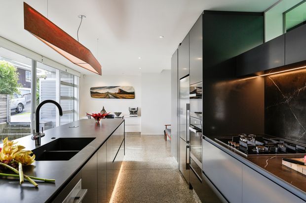 Contemporary galley-style kitchen with a large black island, integrated sink and gooseneck tap, dark cabinetry, black marble-look splashback, and a gas cooktop. Full-height glazing lines one side, while a linear timber feature pendant adds warmth above the island. Polished concrete or terrazzo-like flooring and concealed LED strip lighting create a refined, high-contrast interior.