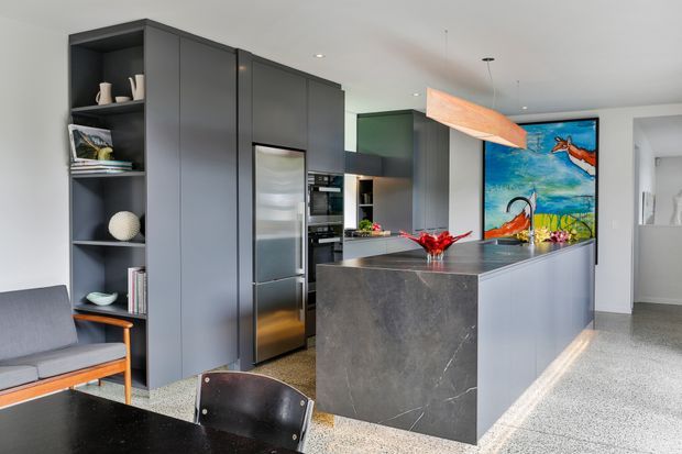 A contemporary open-plan kitchen and dining area with dark grey floor-to-ceiling cabinetry, integrated stainless-steel appliances, a large stone island with waterfall edge, and a suspended timber feature light. Open shelving, terrazzo-style flooring, colourful artwork, and mid-century-inspired seating add visual interest.