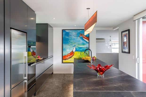 Contemporary open-plan kitchen with dark glossy cabinetry, integrated stainless-steel refrigerator, a large dark stone island with an undermount sink and sculptural red glass bowl, and colourful artwork. A dining or living area with chandelier is visible beyond the kitchen.