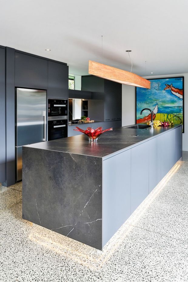 Contemporary kitchen with a large central island, matte dark cabinetry, integrated stainless-steel appliances, a dark veined stone island benchtop and pale blue-grey island cabinetry. The room has a polished terrazzo floor, white ceiling with recessed lighting, a sculptural timber pendant, and a colourful feature artwork at the rear.