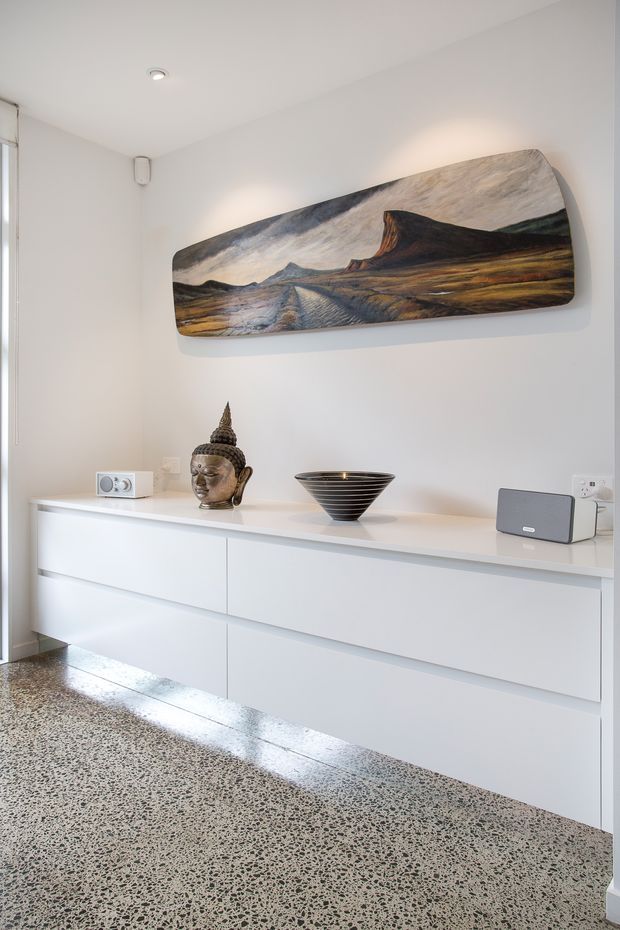 A bright, minimalist interior featuring a long white built-in sideboard with handleless drawers, a polished speckled terrazzo floor, and white walls. Decorative objects including a Buddha sculpture, black bowl, radio, and speaker sit on the sideboard, beneath a large landscape artwork. A tall window or glazed opening is visible at the left.