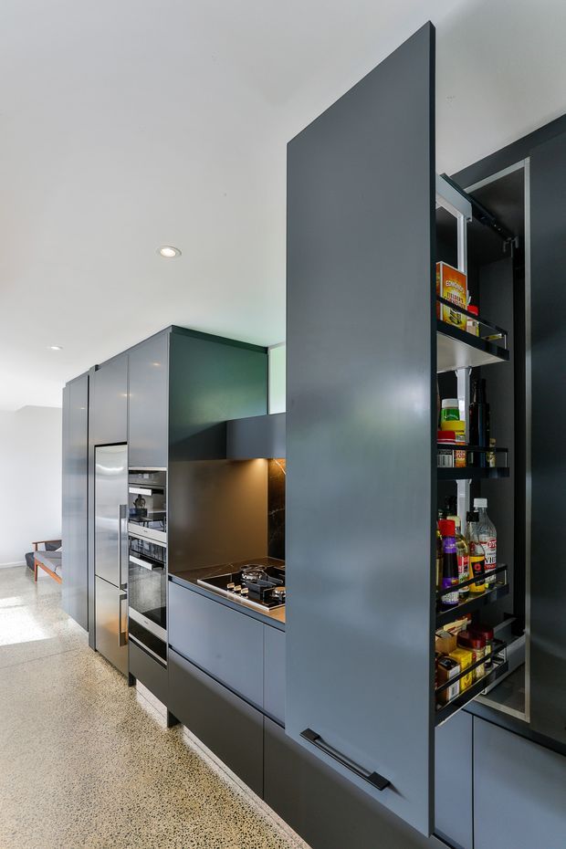 Contemporary galley kitchen with tall matte dark-grey cabinetry, integrated appliances, a concealed pull-out pantry, dark benchtops and a warm metallic splashback. Polished concrete or terrazzo-like flooring and recessed ceiling lights complete the sleek, functional fit-out.