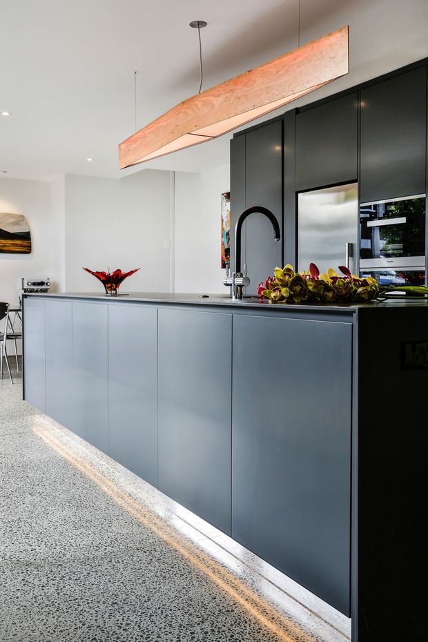 Contemporary kitchen with a long dark island featuring integrated cabinetry, a black gooseneck tap and sink, tall black cabinetry with built-in appliances, and a light grey terrazzo floor. A sculptural timber pendant light hangs above the island, with colourful decorative foliage adding contrast.
