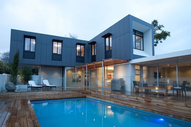 Contemporary multi-level residence with a dark blue-grey panelled upper facade, expansive glazing, projecting box forms, and a timber pergola over the outdoor entertaining area. The house opens onto a timber pool deck surrounding a bright blue swimming pool, with visible outdoor dining and lounge spaces. The image is an exterior architectural view photographed at dusk.