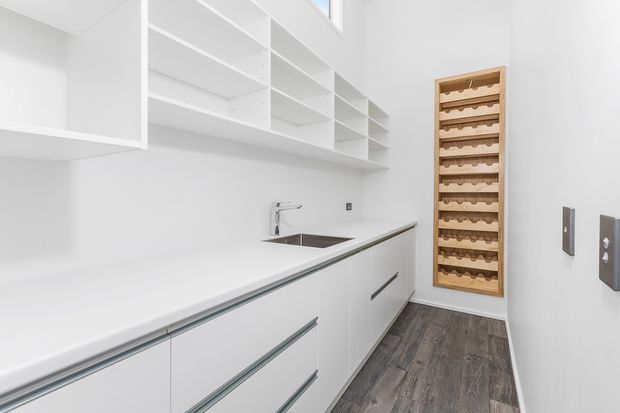 Bright, narrow white walk-in pantry with extensive open shelving, white built-in cabinetry, a long pale benchtop, undermount sink and tap, integrated drawer fronts, dark wood-look flooring, and a built-in wooden wine rack at the far end.