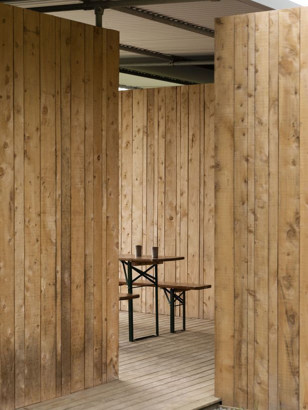 A compact semi-enclosed outdoor dining or entertaining area framed by tall vertical timber plank screens. The space has a timber decking floor, exposed dark metal supports, and a corrugated metal roof, with a simple high table and bench seating visible through the opening. The raw timber and utilitarian structure create a rustic, industrial-leaning atmosphere.