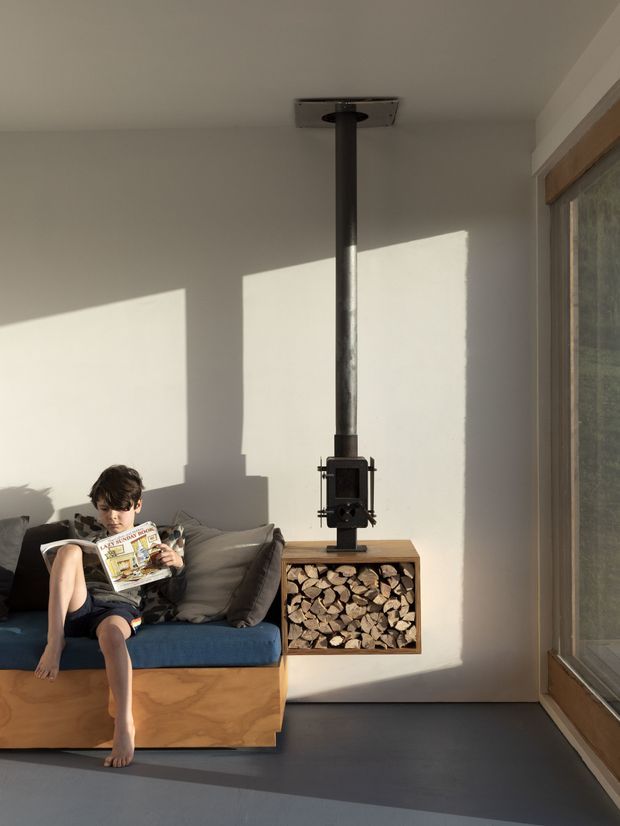 A bright, minimalist living room with a built-in timber-framed sofa and blue upholstery beside a compact black wood-burning stove. The stove has a tall flue extending to the ceiling and sits above an open timber log-storage cabinet filled with firewood. White plaster walls, a dark smooth floor, and large timber-framed glazing create strong late-afternoon shadows across the room.