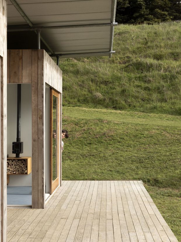 A minimalist small timber-clad dwelling opening onto a broad pale timber deck. The image looks from the covered exterior deck toward a grassy sloping site, with a large glazed timber-framed door and a glimpse of an interior wood-burning stove and firewood storage. The building has a simple corrugated metal canopy supported by slender posts and a restrained natural material palette.