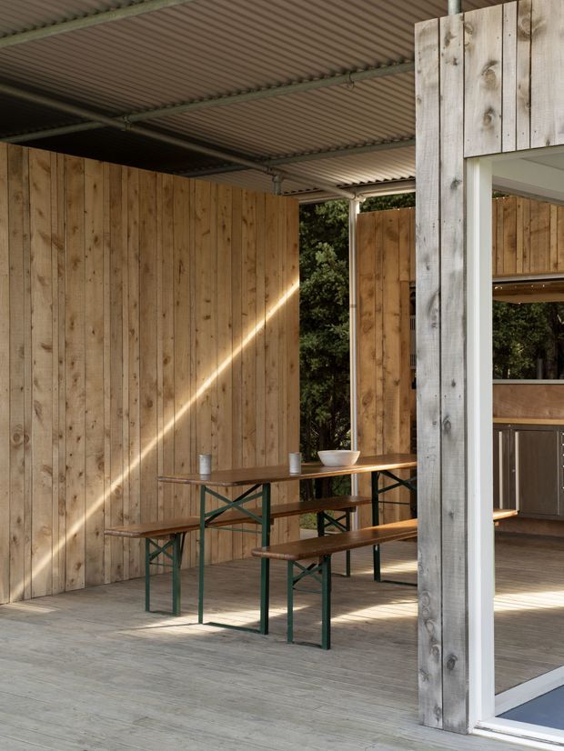 A covered outdoor entertaining area with weathered timber-clad walls, a corrugated metal roof and timber decking. A long timber picnic-style table with matching benches and dark green metal frames occupies the space, while a partially visible built-in outdoor kitchen or bar appears behind a glazed opening on the right. Sunlight filters through the surrounding vegetation, creating a warm, rustic atmosphere.