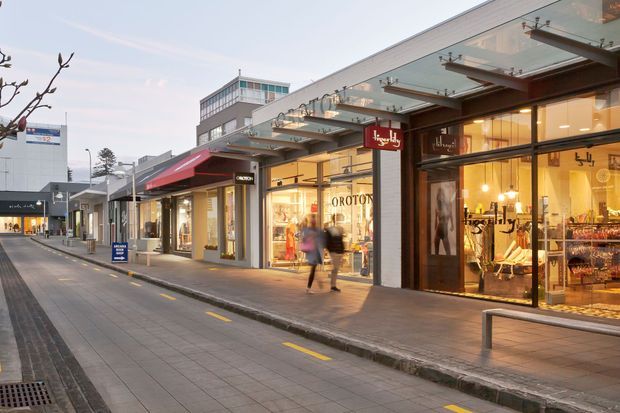 Street-level view of a contemporary low-rise retail shopping precinct with continuous glazed shopfronts, projecting awnings and canopies, illuminated storefronts, pedestrian paving, and a narrow access lane with yellow road markings. Multiple fashion and retail businesses are visible, including Oroton, with warm interior lighting contrasting against the cool early-evening exterior.