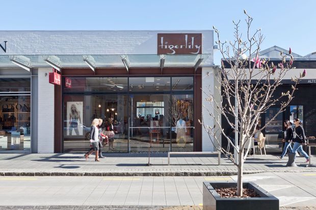 Street-facing retail storefront in a contemporary open-air shopping precinct. The main facade has extensive floor-to-ceiling glazing, a shallow projecting glazed canopy, light rendered or masonry framing, and a dark shop sign. Pedestrians and a street tree are visible in the foreground, with neighbouring retail tenancies extending on both sides.
