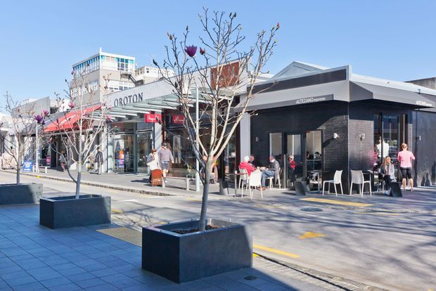 A sunny urban shopping precinct with a modern single-storey black-brick café or commercial tenancy in the foreground, featuring a broad low-pitched roof, dark facade, outdoor tables and chairs, and large glazed openings. Adjacent retail storefronts and multi-storey commercial buildings line the paved pedestrian streetscape, with young deciduous trees in large dark planters.