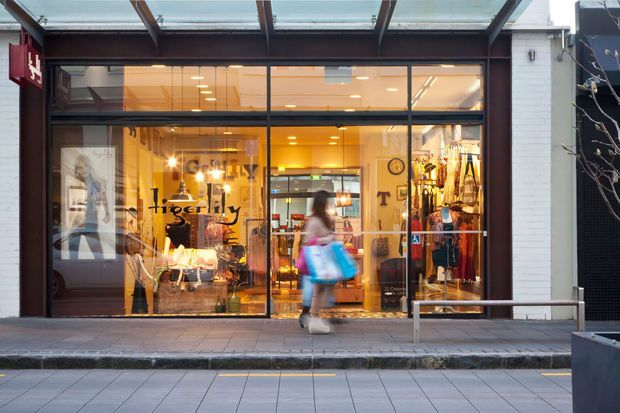 Street-level photograph of a boutique retail shopfront with a fully glazed facade, dark metal framing, warm illuminated interior, clothing displays and signage. A covered canopy projects over the footpath, with a pedestrian passing in front.