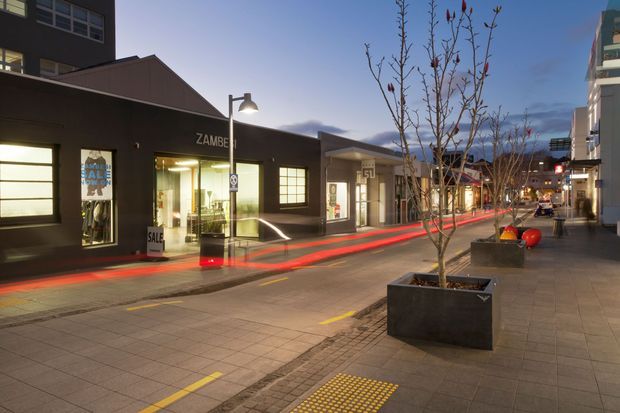 Twilight streetscape in a contemporary urban commercial precinct, with low-rise dark-clad shopfronts, illuminated retail windows, pedestrian paving, street trees in large planters, bollards, and modern apartment or office buildings along the right side. Red linear lighting or illuminated street edging creates a strong visual accent, while tactile paving and marked pedestrian areas are visible in the foreground.
