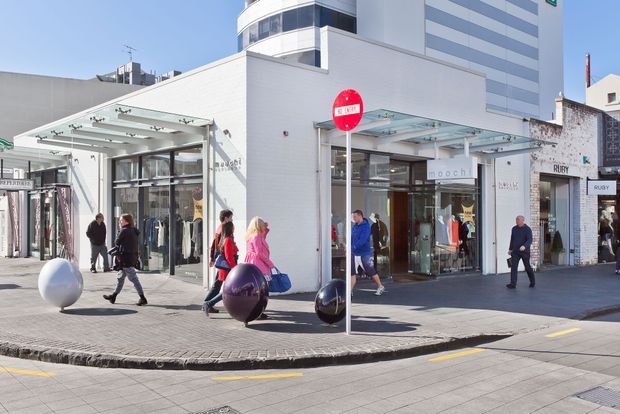 Street-level view of a contemporary urban retail frontage on a paved pedestrian corner. The low white-brick commercial building has large glazed shopfronts, projecting glass-and-metal entrance canopies, clothing and fashion retail displays, and a taller modern building rising behind it. Pedestrians and sculptural spherical street furniture occupy the foreground, with a visible no-entry sign and adjacent shop signage.