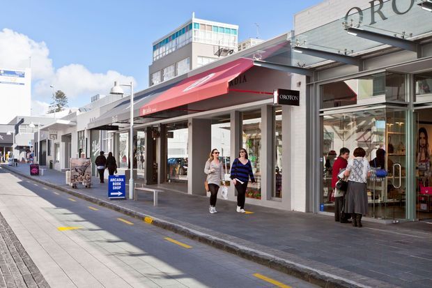 Street-level view of a bright urban retail shopping strip with modern low-rise storefronts, extensive glazing, shop awnings, pedestrian paving, signage, and several people along the footpath. The image primarily documents the commercial streetscape and building facades.
