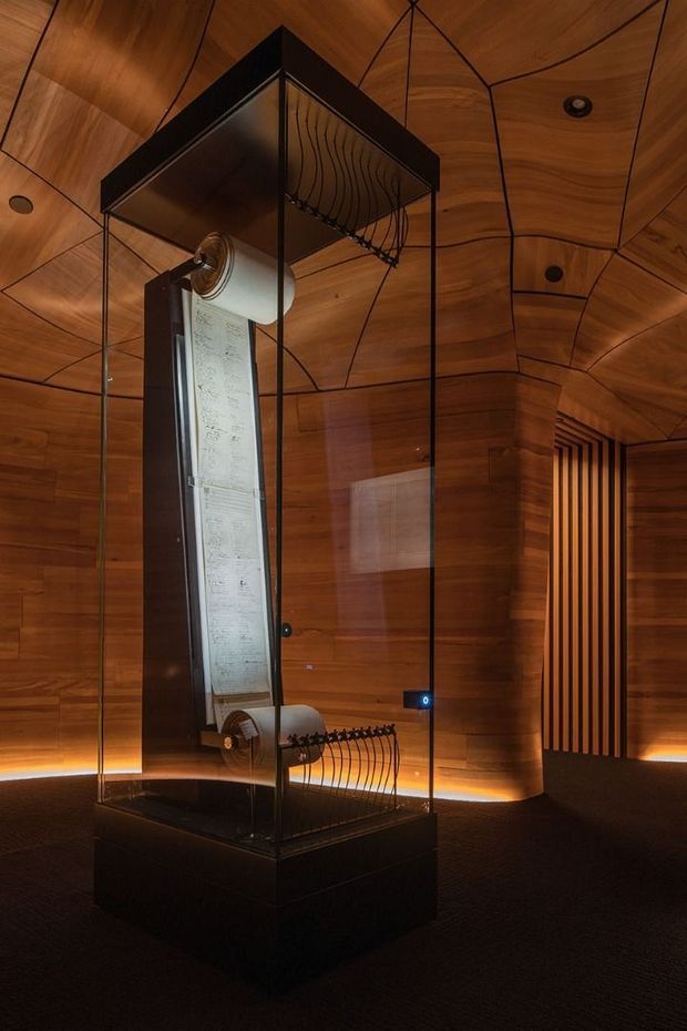 A dimly lit, museum-like interior featuring a tall glass display case containing an illuminated scroll or document. The room is enveloped in warm timber wall and ceiling panels with dark linear joints, concealed perimeter lighting, dark carpet, and a vertical slatted timber feature wall. The display case has a dark plinth and decorative curved metal supports, creating a dramatic, refined exhibition atmosphere.