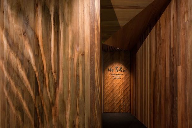 A narrow, dimly lit interior passage lined almost entirely with richly patterned vertical timber wall panelling. The corridor leads toward a darker end wall featuring a small graphic or text panel, with timber also visible on the ceiling and a strong diagonal structural or ceiling element overhead. The composition emphasises warm natural wood grain, shadow, and an immersive enclosed atmosphere.