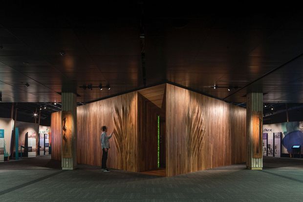 A dark, immersive exhibition or commercial interior featuring a sculptural, angular timber-clad pavilion at the centre. Warmly illuminated wood walls and a narrow green-lit entrance contrast with a black ceiling, track lighting, patterned carpet, display panels, and structural columns around the space.