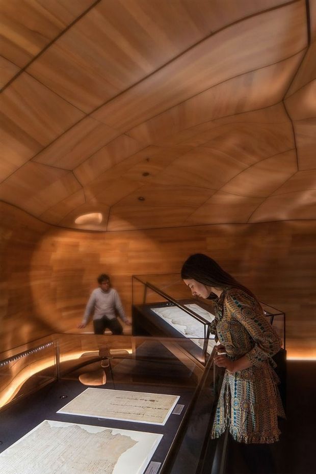 An immersive exhibition or museum interior with a sculptural, organically curved timber-lined ceiling and walls. Dark display tables with illuminated glass cases present documents and maps, while visitors examine the exhibits. Warm amber lighting and continuous wood panelling create an intimate, enveloping atmosphere.