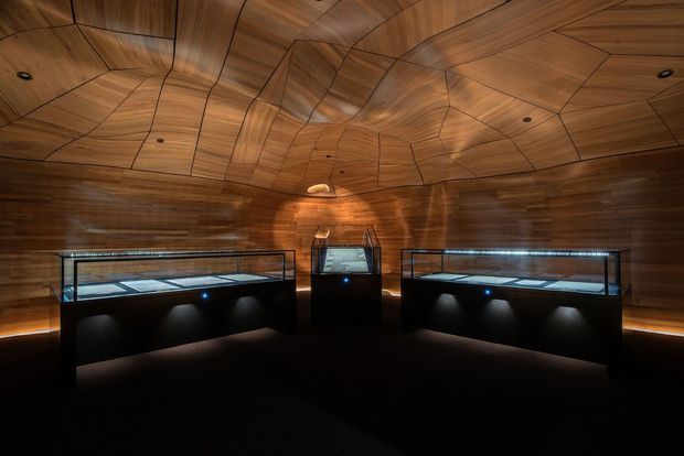 A dark, immersive exhibition or gallery interior with richly finished curved timber wall and ceiling panels forming a sculptural, faceted enclosure. Three illuminated glass display cases with dark built-in bases occupy the room, while discreet recessed lighting and concealed linear illumination create a dramatic museum-like atmosphere.