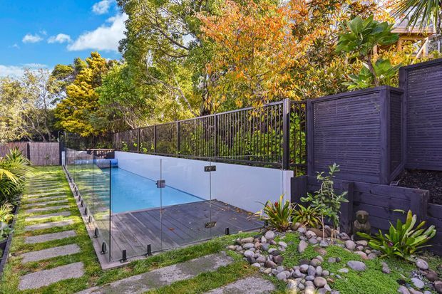 A landscaped outdoor pool area with a narrow rectangular swimming pool enclosed by frameless glass fencing. Dark timber decking, white rendered retaining walls, black metal fencing, timber privacy screening, stepping-stone paths, pebble beds, and lush tropical planting create a contemporary garden setting.